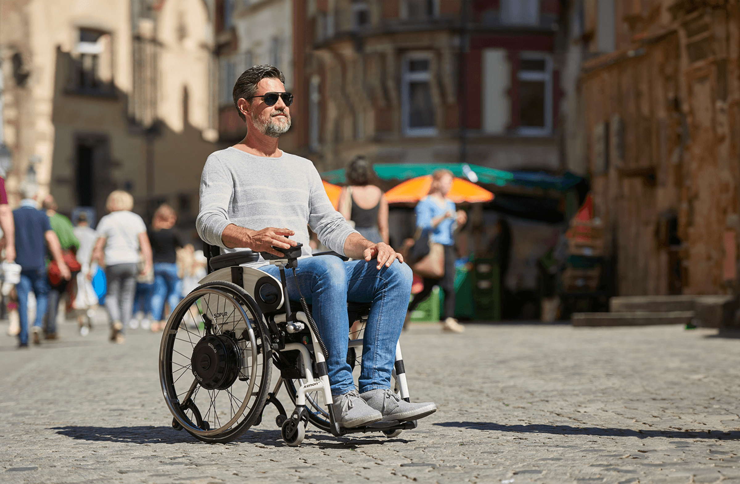 The picture shows a middle-aged man, wearing sunglasses, driving through a pedestrian zone on a summer day in his wheelchair with an auxiliary drive for self-propelled driving. The add-on drive is black and installed in the wheels as a wheel hub drive.