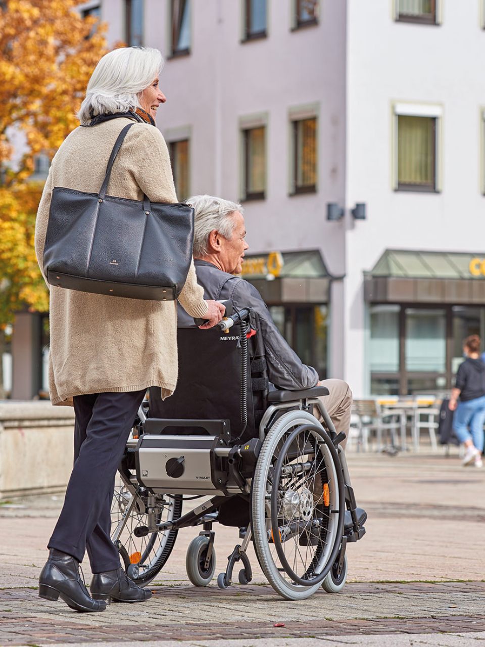 The picture shows a woman driving an elderly man in a wheelchair through a city centre with the help of the pushing and braking aid V-MAX2. This picture should stand for the information contribution on the topic of aids in the nursing home. 