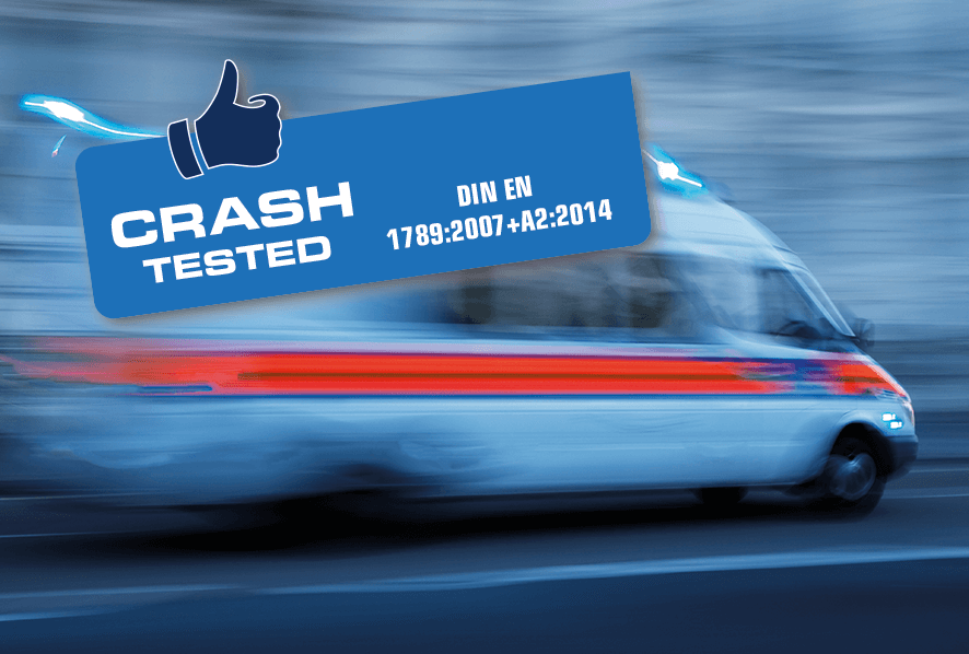 Das Bild zeigt einen Krankentransportwagen im Vorbeifahren. Aufgrund der Geschwindigkeit verschwimmt das Bild. Im Vordergrund befindet sich ein Logo, das besagt, dass der CR23 Crash getestet nach DIN EN 1789:2007-A2:2014 ist und somit auch über ein Halterungssystem im Fahrzeug mitgeführt werden darf.