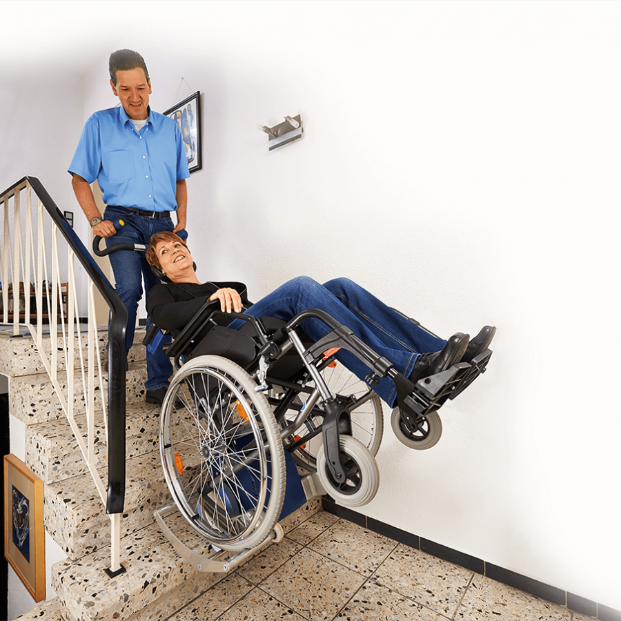 Das Bild zeigt einen Mann mittleren Alters, der eine Frau im Rollstuhl über Treppen innerhalb eines Hauses befördert. Der Rollstuhl wurde hierzu auf die Aufnahmeschuhe des Treppensteiger s-max in Kombination mit der Universalaufnahme SDM7 geschoben.