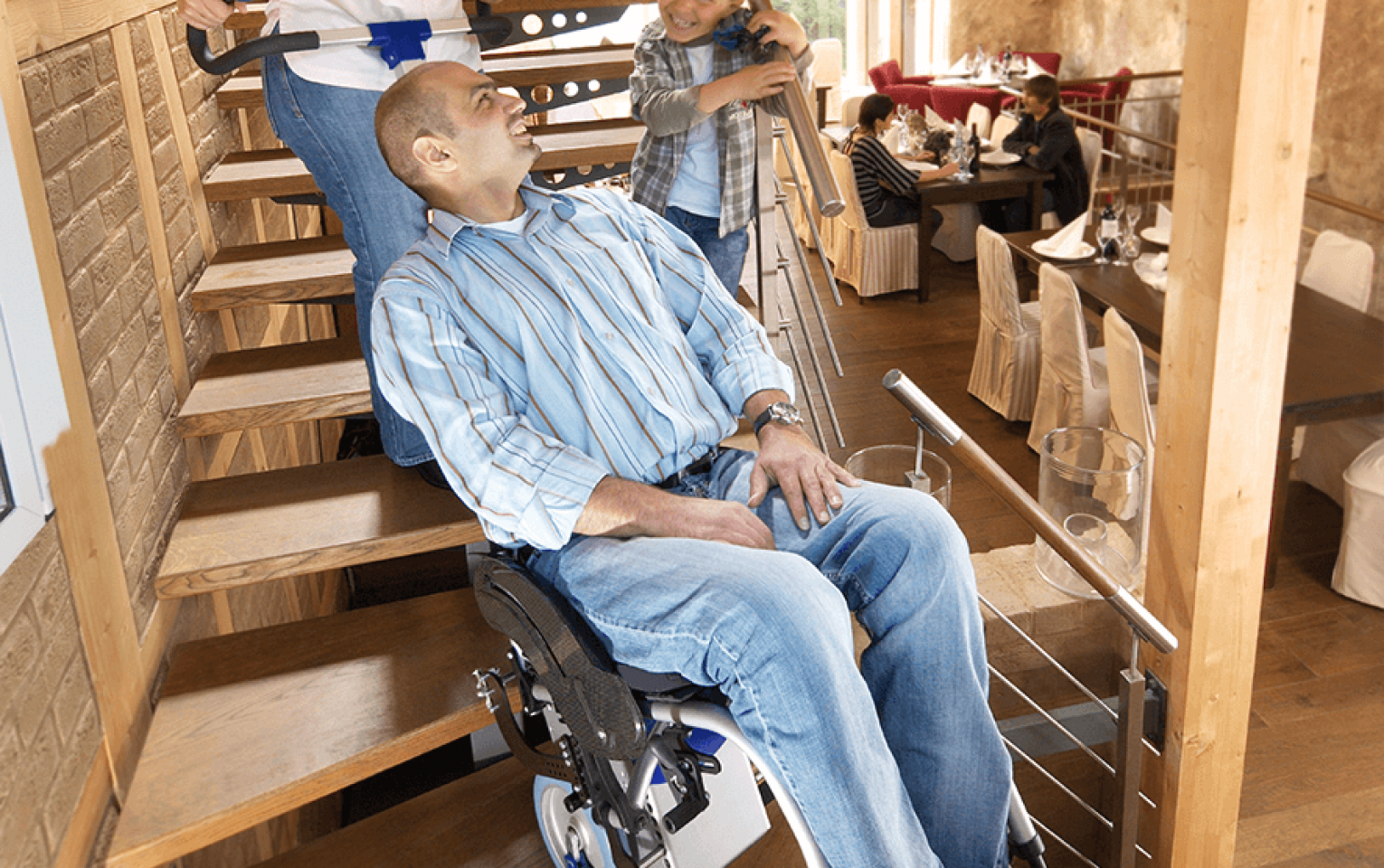 Das Bildzeigt einen Mann, der in seinem Rollstuhl sitzt und von einer Frau mit Hilfe eines angebauten Treppensteigers über eine Treppe transportiert wird.