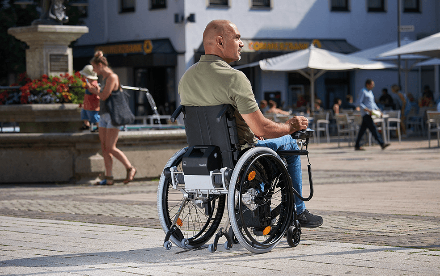 The picture shows a man in a wheelchair who controls his wheelchair himself using a joystick control unit. The wheelchair is fitted with an auxiliary drive from AAT - a MAX-E in the Black Edition.