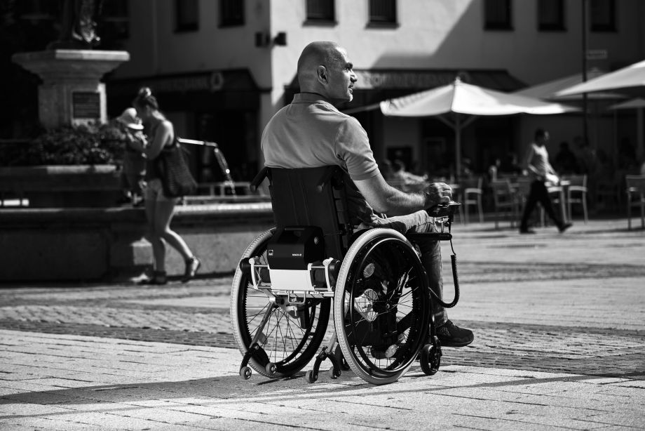 The picture shows a middle-aged man driving his MAX-E add-on drive through the city center. A café and a large fountain can be seen in the background. He is driving on cobblestones of different sizes.