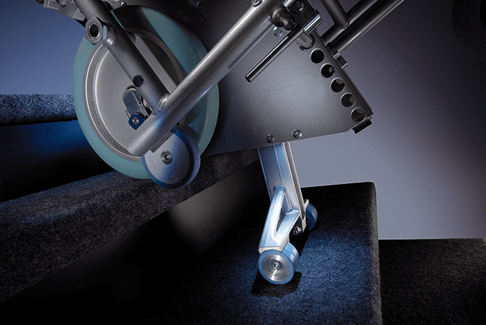 Das Bild zeigt den Steigvorgang der Treppenhilfe s-max. Der Steigfuß ist ausgefahren und setzt bereits auf der unteren Treppenstufe auf. Die Sicherheitsbremsen, welche am großen Laufrad befestigt sind, setzen am Stufenrand auf und schützen so vor einem abwärtsrollen. Die LED-Leuchte erhellt die Treppenstufen.