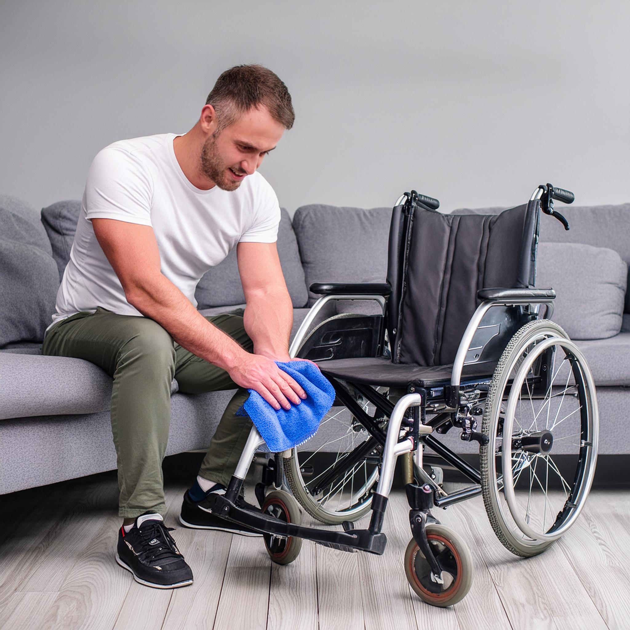 Das Bild zeigt einen jungen Mann, der im Wohnzimmer auf dem Sofa sitzt und seinen Rollstuhl mit einem Tuch reinigt. 