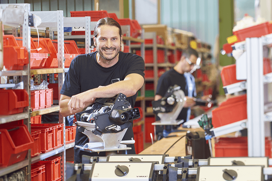 Das Bild zeigt einen Mitarbeiter der Firma AAT Alber Antriebstechnik an seinem Arbeitsplatz in der Produktion. Er lächelt freundlich in die Kamera und hat seinen Arm auf eine Schiebe- und Bremshilfe V-MAX² gelegt, welche er gerade zusammenbaut.