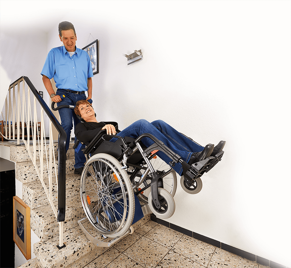 Das Bild zeigt einen Mann mittleren Alters, der eine Frau im Rollstuhl über Treppen innerhalb eines Hauses befördert. Der Rollstuhl wurde hierzu auf die Aufnahmeschuhe des Treppensteiger s-max in Kombination mit der Universalaufnahme SDM7 geschoben. 