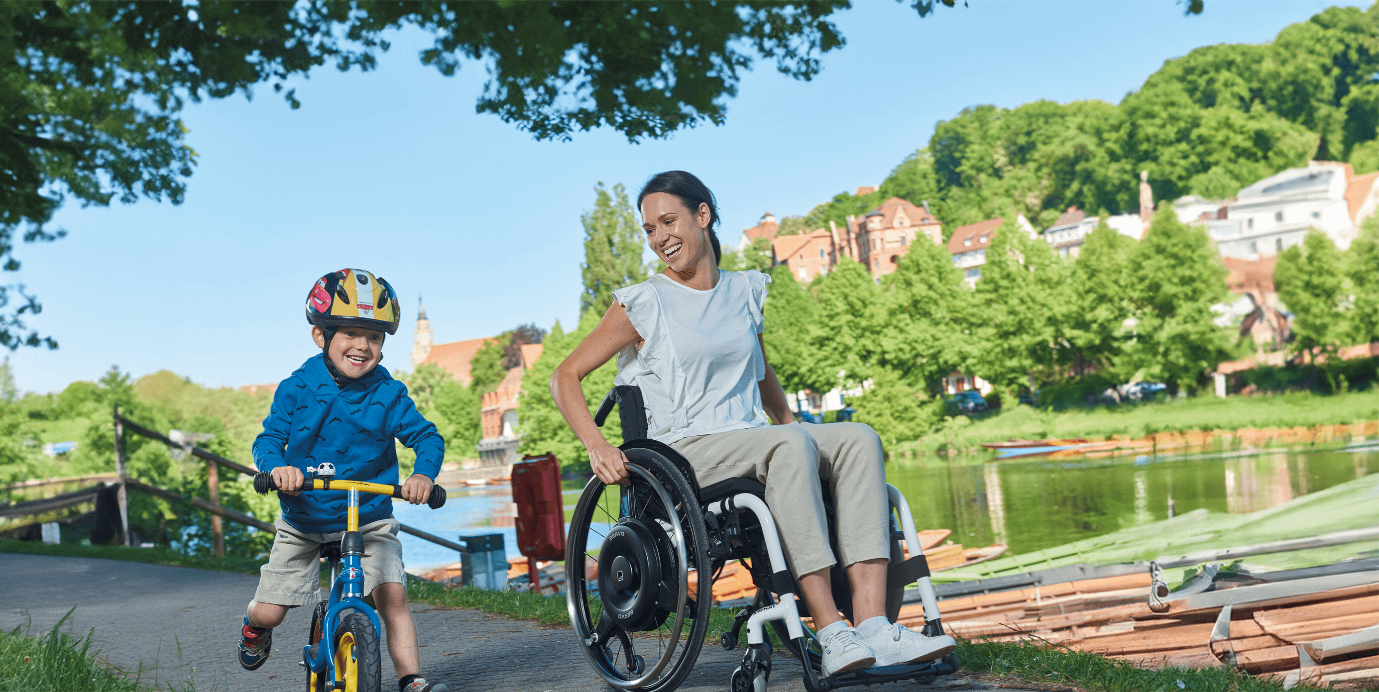 A young woman in a wheelchair uses a black wheel hub drive for remaining power and drives along a river with her son on a city scooter.