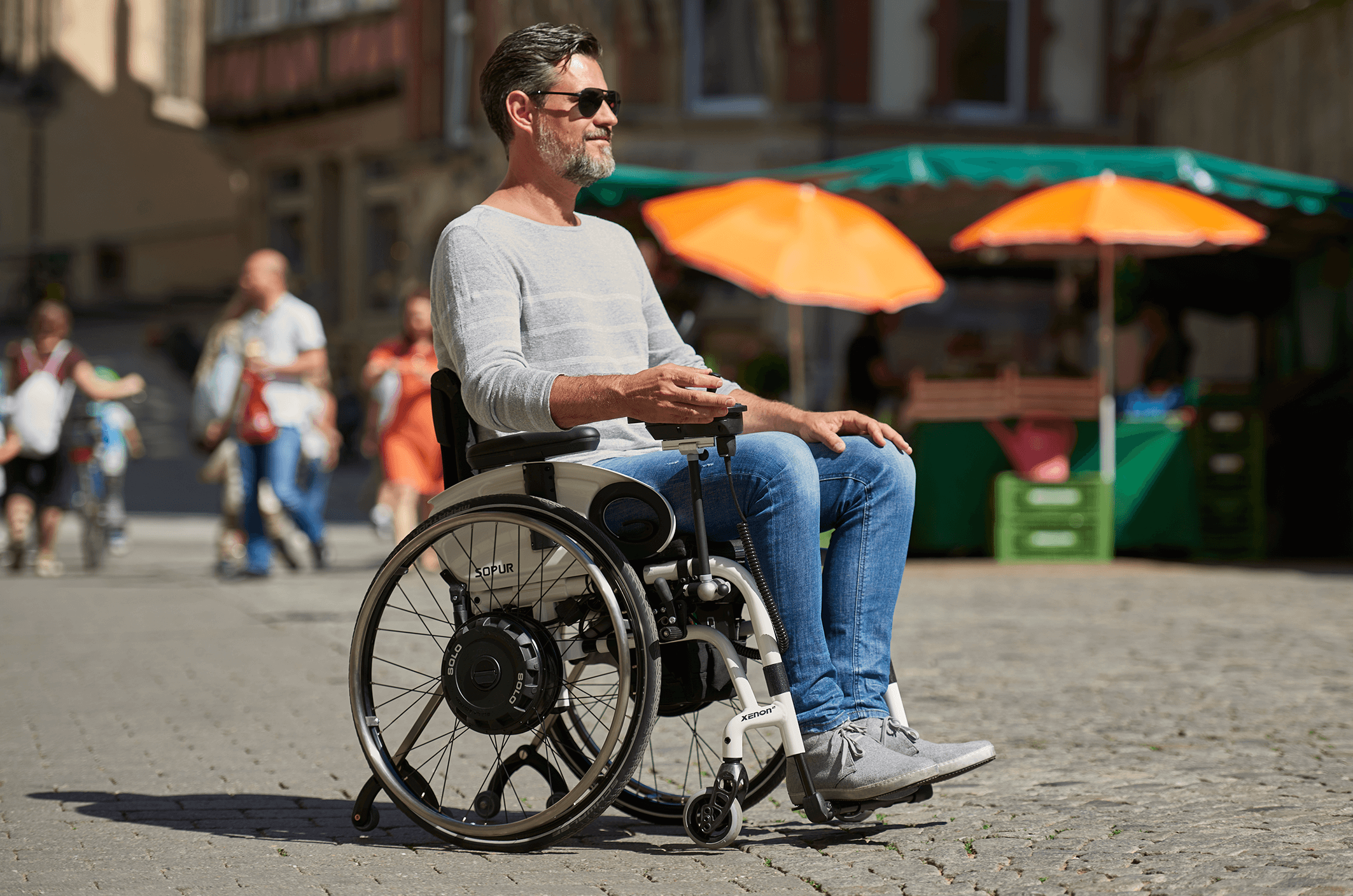 Das Bild zeigt einen Mann mittleren Alters, der an einem sonnigen Tag mit seinem elektrisch betriebenen Rollstuhl mit Zusatzantrieb in der Stadt unterwegs ist. 