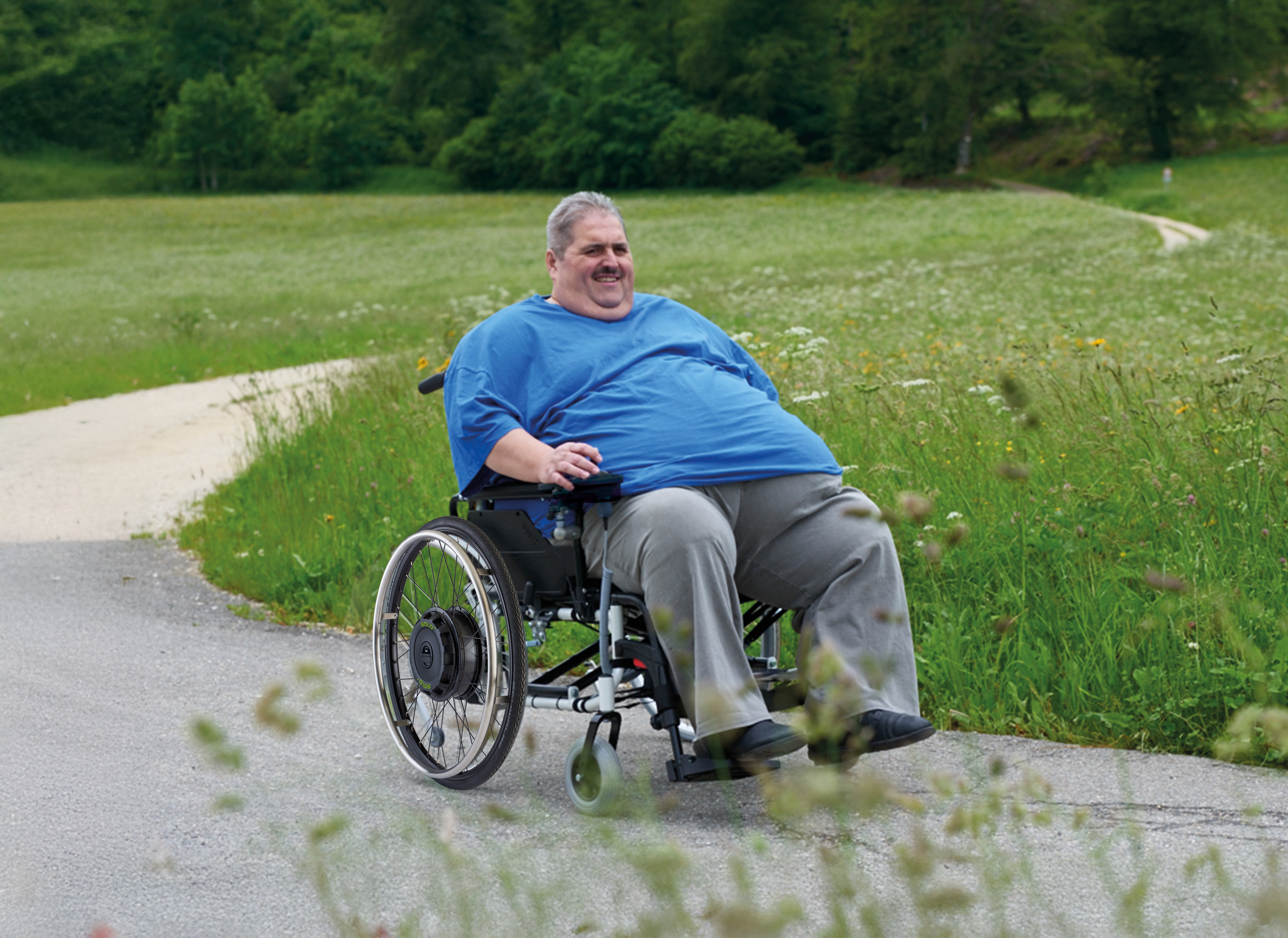 The picture shows a middle-aged man with a high body weight. He is sitting in an XXL wheelchair, which he is steering along a country lane using a SOLO+ wheelchair add-on drive with a control unit. A forest edge and a lot of green meadow are visible in the background.