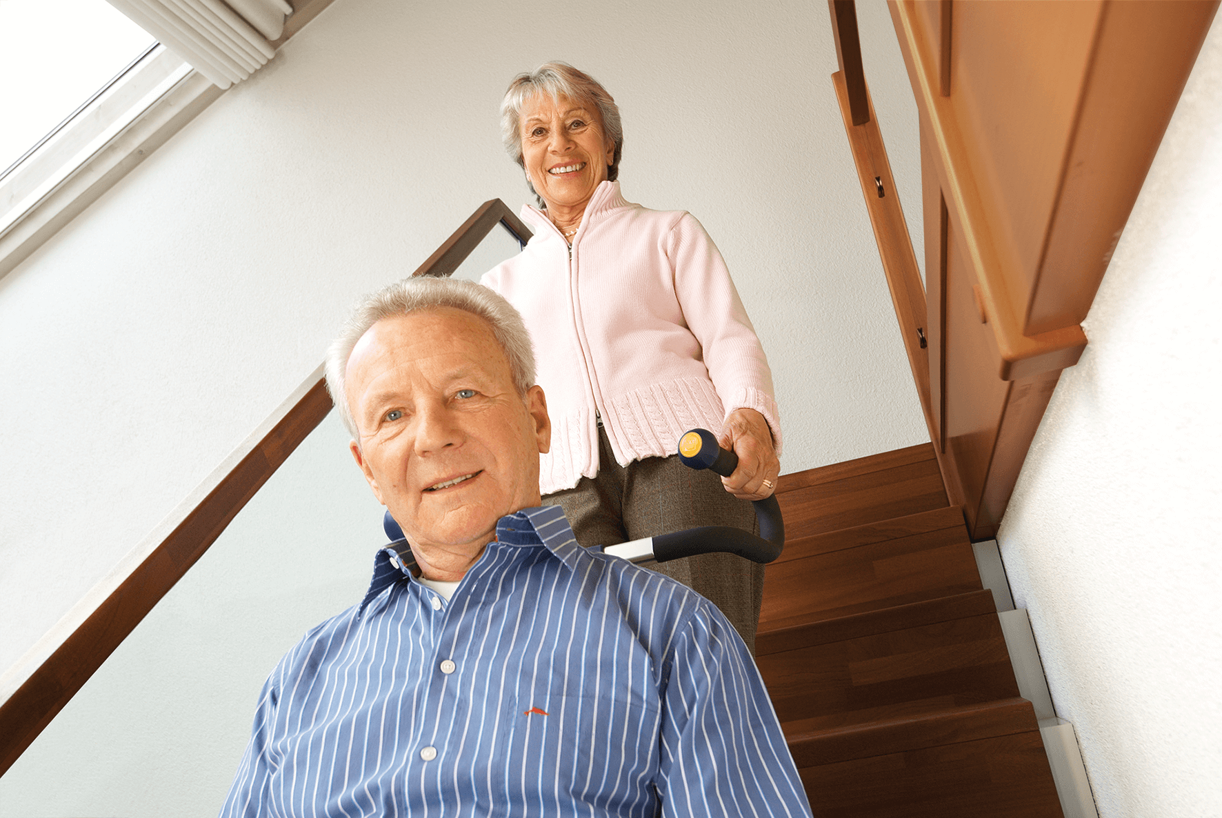 Das Bild zeigt einen Mann mittleren Alters, der von einer Frau in seinem Rollstuhl mithilfe eines Treppensteigers die Treppen hinaufbefördert wird. Beide lächeln in die Kamera.