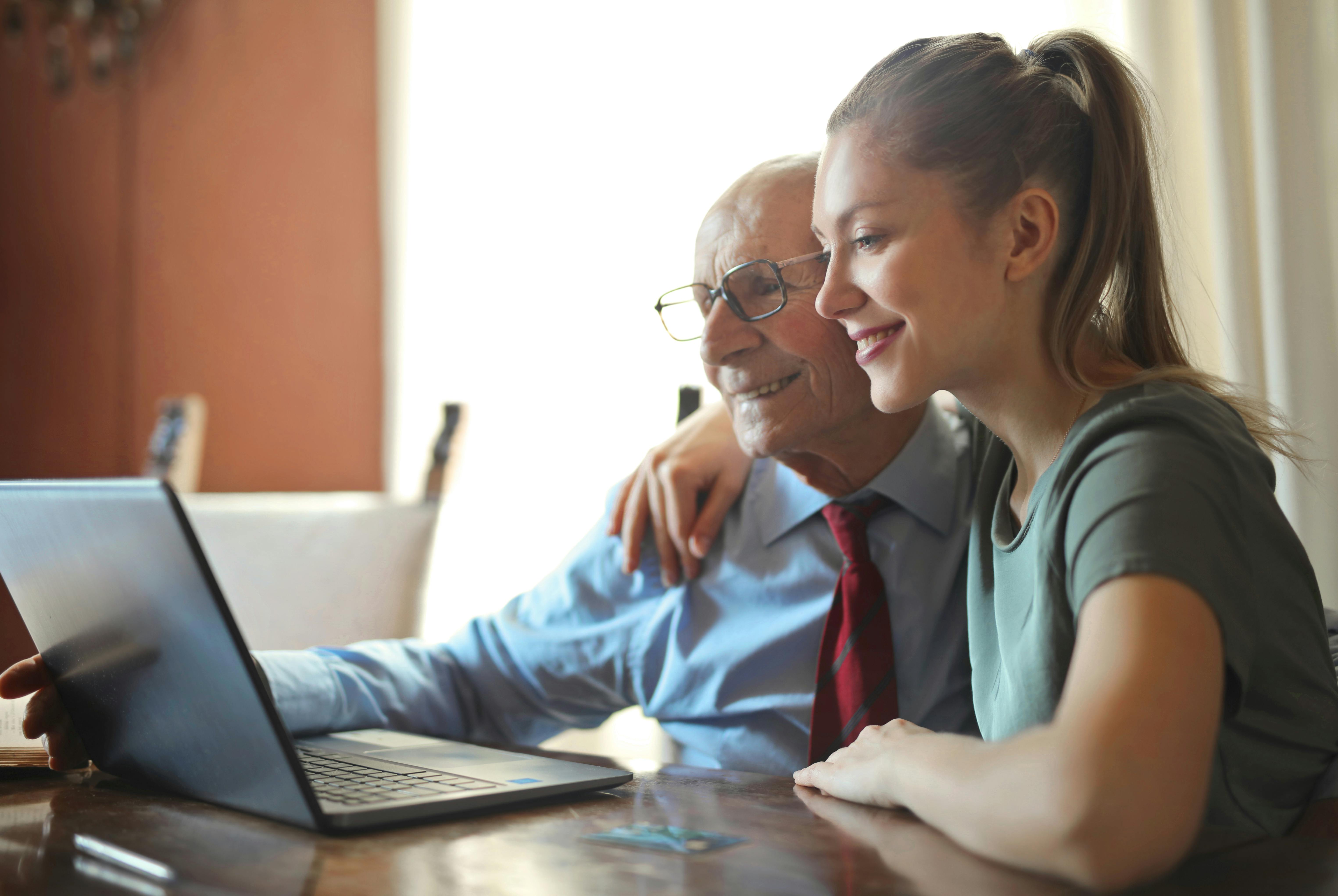 Eine junge Frau unterstützt einen älteren Mann am Laptop, während sie gemeinsam den Widerspruch gegen die Ablehnung eines Hilfsmittelantrags vorbereiten.