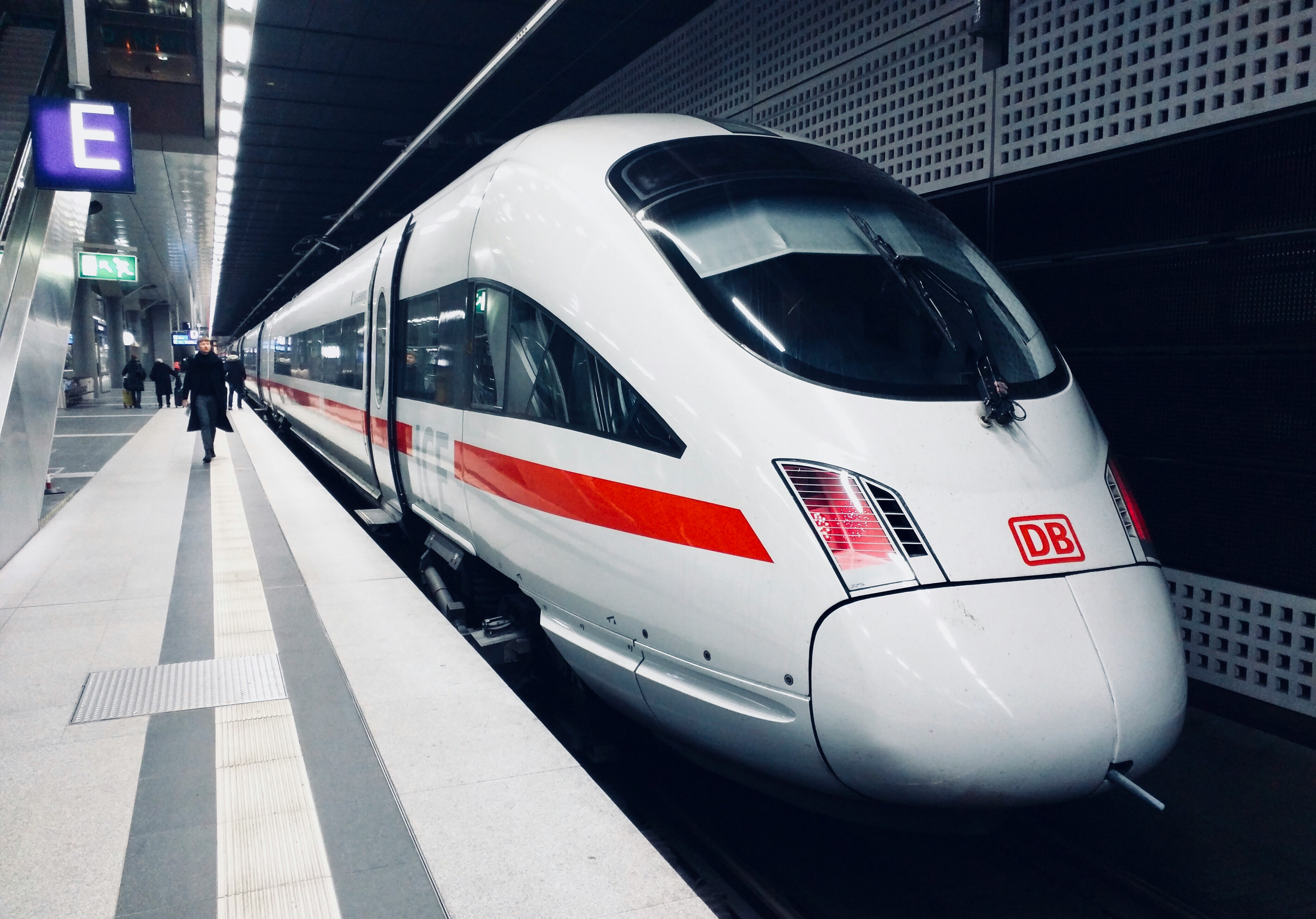 Das Bild zeigt einen modernen Bahnhof in Deutschland, an dem ein Zug der DB am Gleis steht. Im Hintergrund befinden sich einige Fahrgäste.