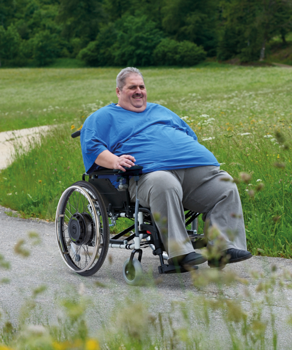 The picture shows a middle-aged man with a high body weight. He is sitting in an XXL wheelchair, which he is steering along a country lane using a SOLO+ wheelchair add-on drive with a control unit. The edge of a forest and a green meadow can be seen in the background. 