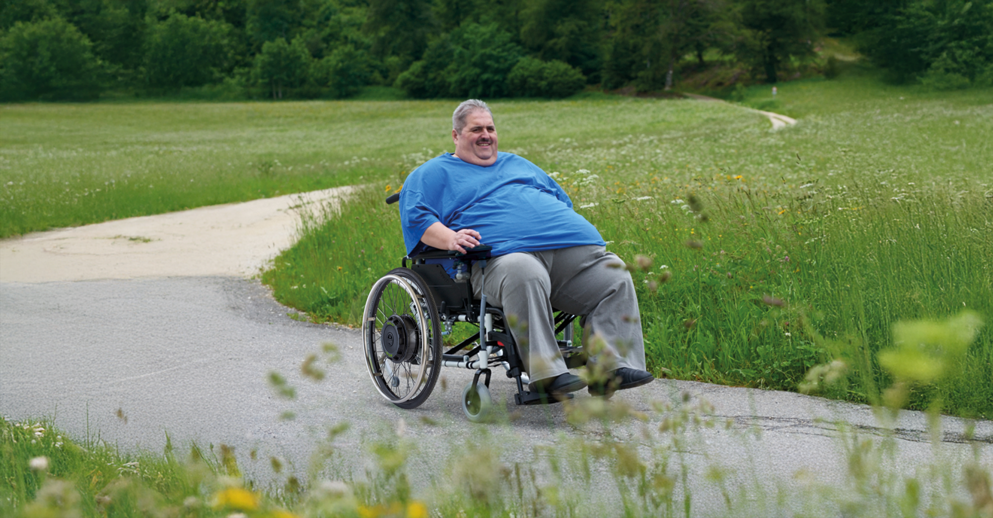 The picture shows a middle-aged man with a high body weight. He is sitting in an XXL wheelchair, which he is steering along a country lane using a SOLO+ wheelchair add-on drive with a control unit. The edge of a forest and a green meadow can be seen in the background.