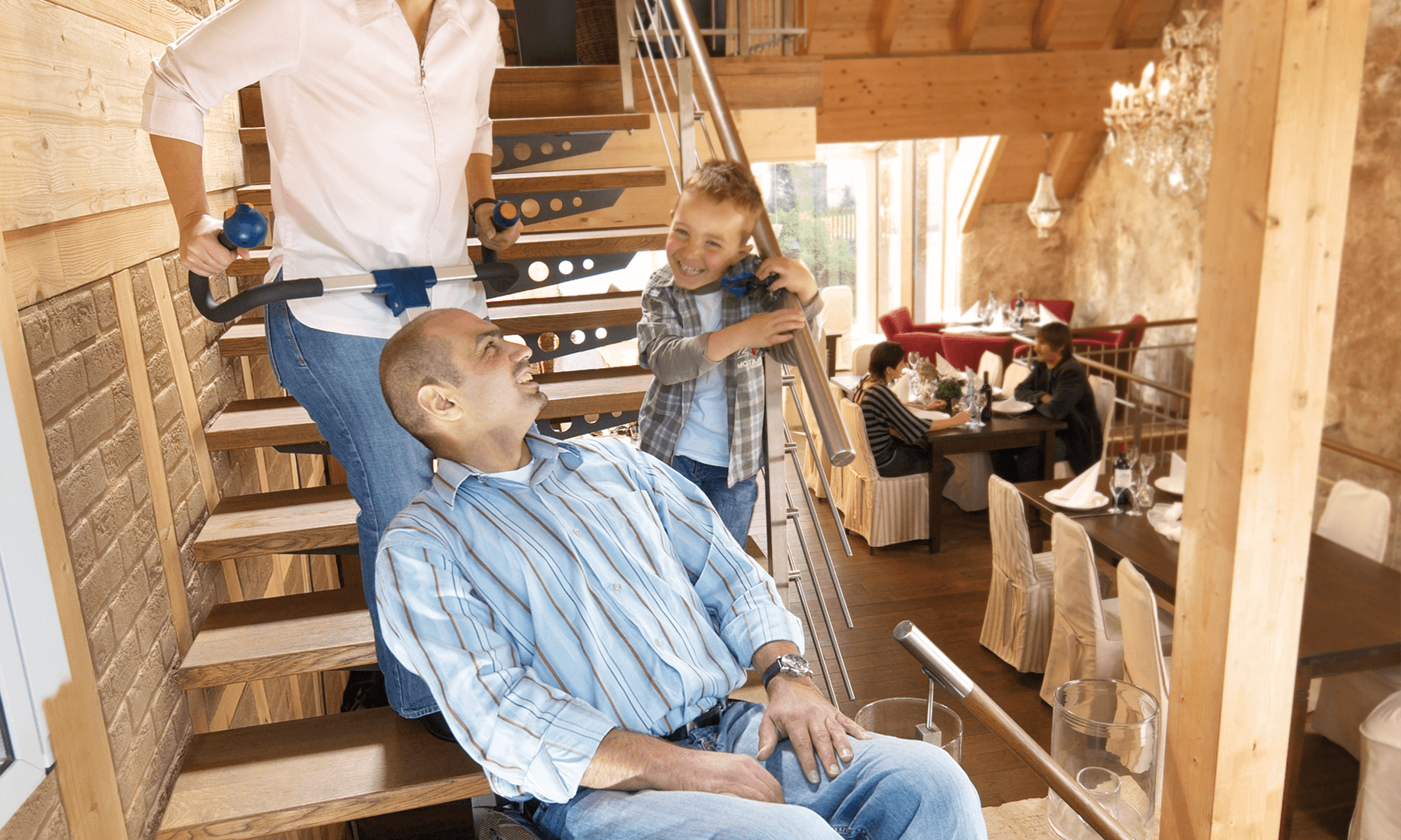 Das Bild zeigt einen Mann mittleren Alters, der von einer Frau in seinem Rollstuhl mithilfe eines Treppensteigers die Treppen hinaufbefördert wird. Neben ihm auf der Treppe steht ein kleiner Junge und lacht ihn an. 