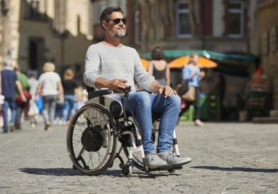 Das Bild zeigt einen Mann mit Sonnenbrille, der an einem sonnigen Tag mit seinem Rollstuhl durch die Stadt fährt. Er nutzt einen Rollstuhl-Zusatzantrieb mit Joystick.