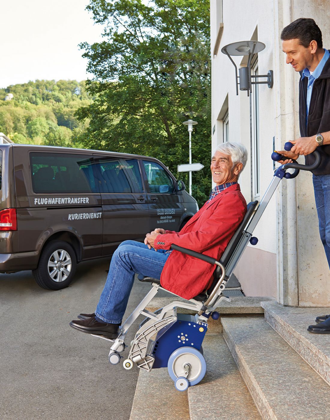 Das Bild zeigt einen Mann mittleren Alters, der einen älteren Mann, der in einem Treppensteiger mit integrierter Sitzeinheit sitzt, die Außentreppe eines Hauses hinunterbefördert. Im Hintergrund steht ein Fahrzeug für den Personentransport / Flughafentransport. 