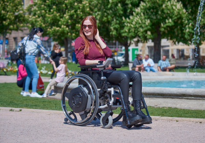 Das Bild zeigt eine junge Frau, die mit ihrem manuellen Rollstuhl inklusive dem neuesten Zusatzantrieb fürü Rollstühle SOLO², in einem belebten Park unterwegs ist.