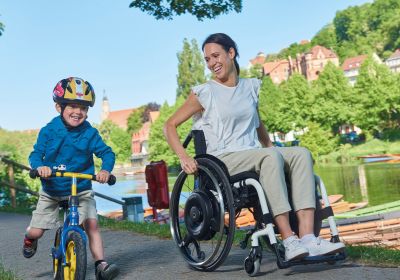 Das Bild zeigt eine Frau im Rollstuhl und ihr Kind mit einem Laufrad. Beide machen einen Spaziergang an einem Fluss. Die Frau nutzt einen Antrieb, der ihre Restkraft unterstützt.