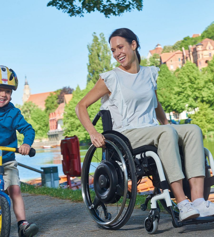 The picture shows a middle-aged man, wearing sunglasses, driving through a pedestrian zone on a summer day in his wheelchair with an auxiliary drive for self-propelled driving. The add-on drive is black and installed in the wheels as a wheel hub drive.