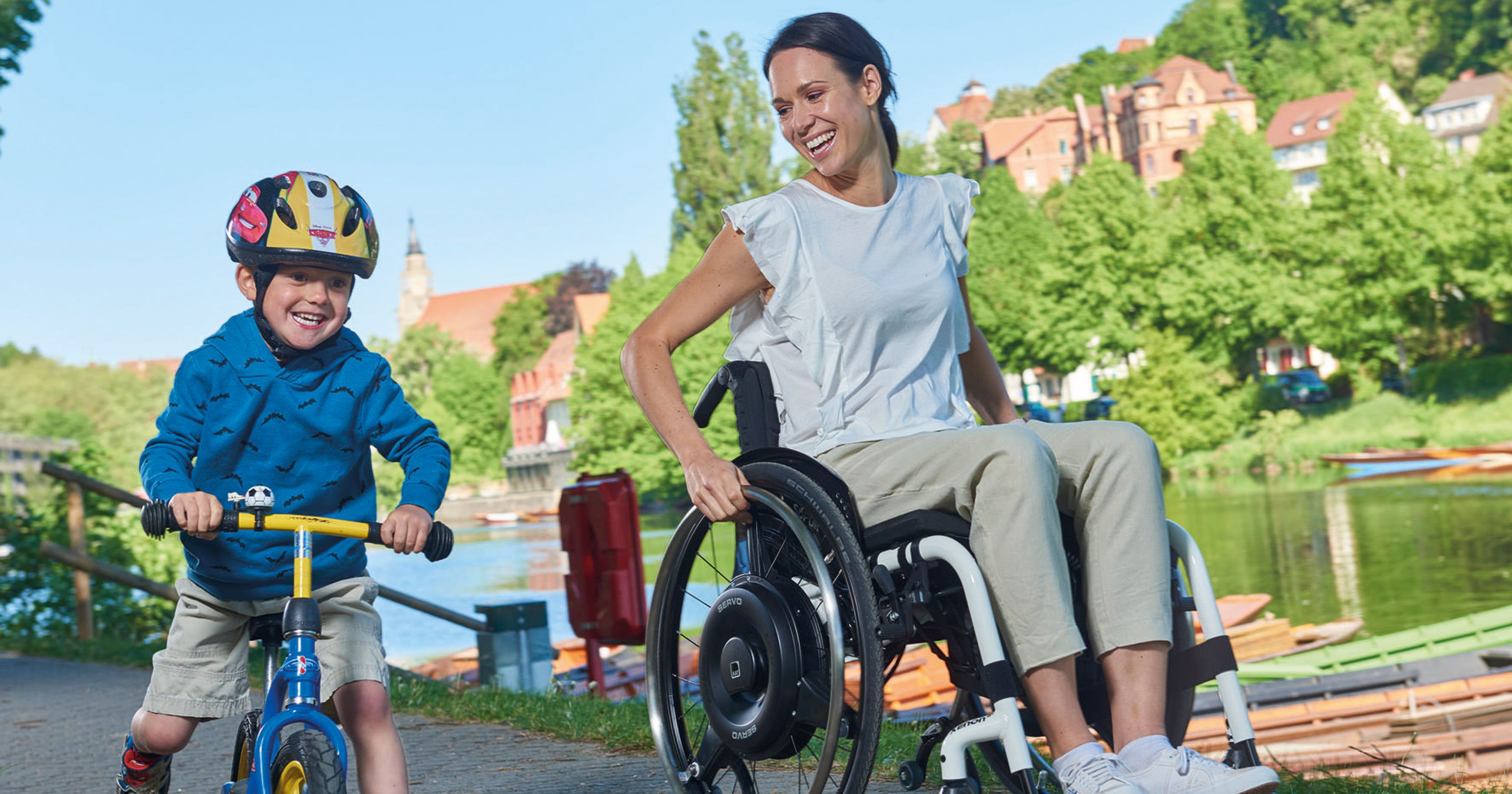 Eine junge Frau im Rollstuhl nutzt einen schwarzen Radnabenantrieb zur Restkraftverstärkung und fährt mit ihrem Sohn auf einem Cityroller an einem Fluss entlang.