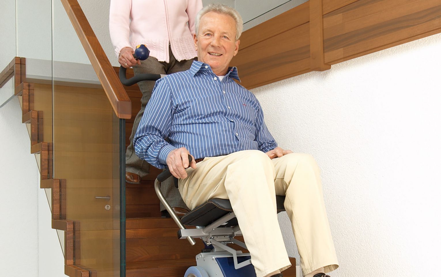 Das Bildzeigt einen Mann, der in seinem Rollstuhl sitzt und von einer Frau mit Hilfe eines angebauten Treppensteigers über eine Treppe transportiert wird. 