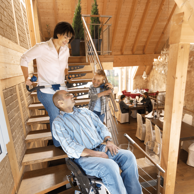 Das Bild zeigt einen Mann mittleren Alters, der von einer Frau in seinem Rollstuhl mithilfe eines Treppensteigers die Treppen hinaufbefördert wird. Neben ihm auf der Treppe steht ein kleiner Junge und lacht ihn an. 