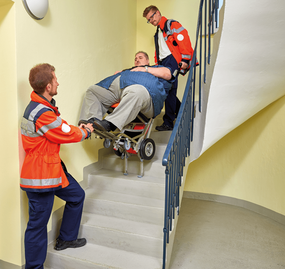 Das Bild zeigt zwei männliche Mitarbeiter eines Rettungsdienstes, die einen sehr adipösen Mann mit über 200 kg Personengewicht mithilfe des Treppensteiger CR23 die Treppe hinauf oder hinab befördern. Ein Mitarbeiter bedient dabei den Treppensteiger und der andere Mitarbeiter führt über den Unterstützungsgriff mit. 