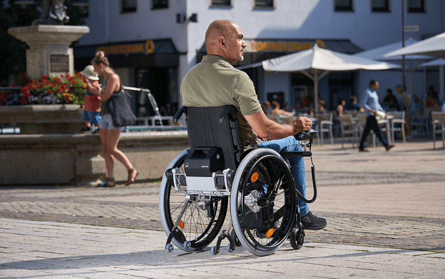 Das Bild zeigt einen Mann in einem Rollstuhl, welcher über ein Bediengerät mit Joystick seinen Rollstuhl selbst steuert. Am Rollstuhl befindet sich ein Zusatzantrieb der Firma AAT - ein MAX-E in der Black Edition. 