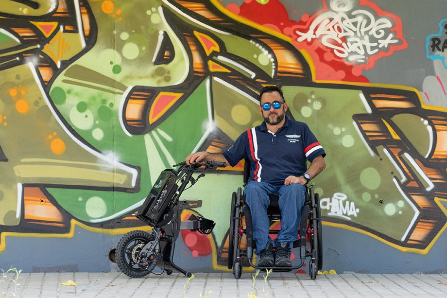 Das Bild zeigt einen Mann mit Sonnenbrille, der mit einem Zuggerät MINI 2 von BATEC, welches er abgebaut und neben seinem Rollstuhl platziert hat, vor einer bunten Wand mit Graffiti sitzt. 