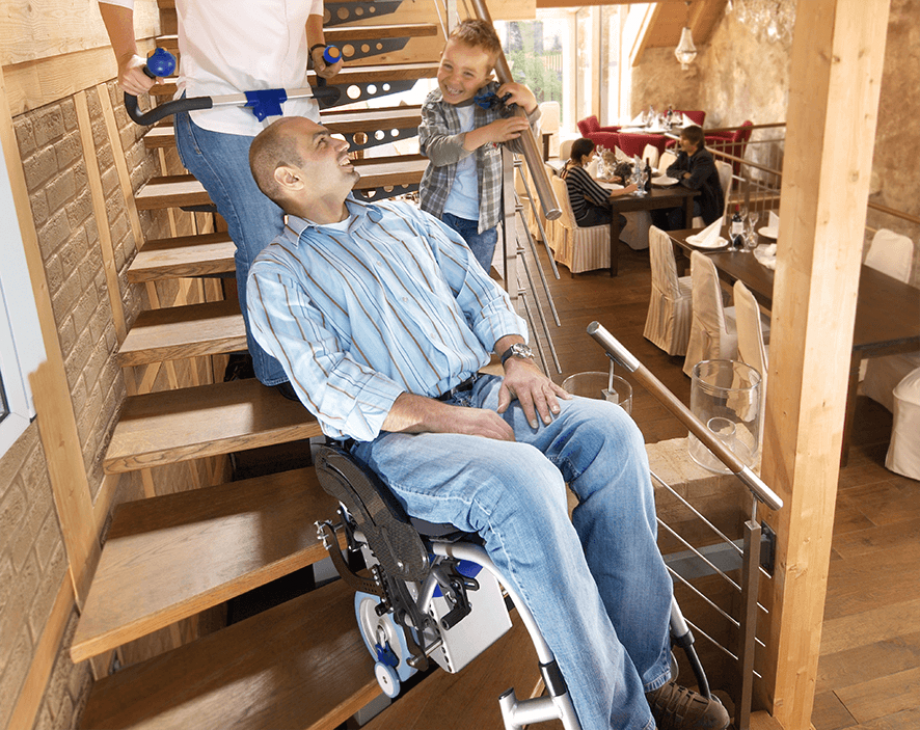 Das Bild zeigt einen Mann mittleren Alters, der von einer Frau in seinem Rollstuhl mithilfe eines Treppensteigers die Treppen hinaufbefördert wird. Neben ihm auf der Treppe steht ein kleiner Junge und lacht ihn an.