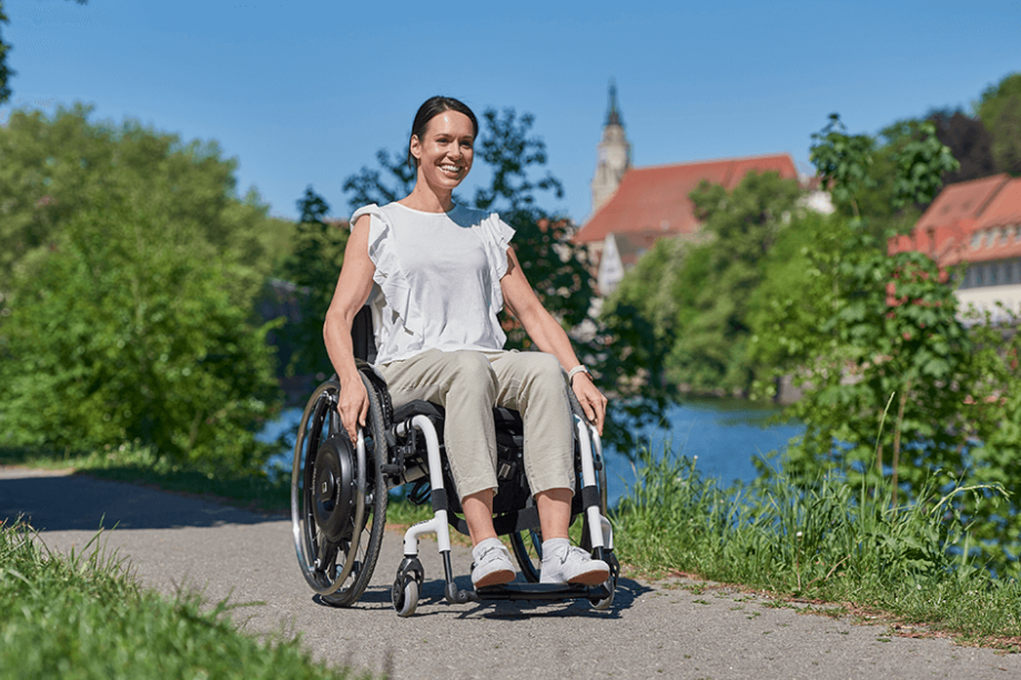 Auf dem Bild ist eine lächelnde Frau mittleren Alters zu sehen, die in einem weißen Rollstuhl mit schwarzem Rollstuhlantrieb zur Restkraftverstärkung lächelnd in Richtung Kamera fährt. Im Hintergrund ist ein Fluss, grüne Bäume und eine Stadt zu sehen.