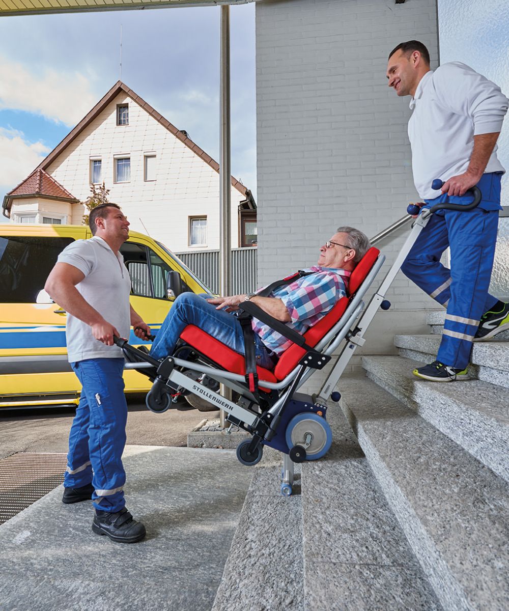 Das Bild zeigt zwei männliche Mitarbeiter eines Krankenfahrdienstes, die einen älteren Mann in einem Tragestuhl mit angebrachtem Treppensteiger eine Treppe hinaufbefördern. Im Hintergrund steht das Krankenfahrdienstfahrzeug. 