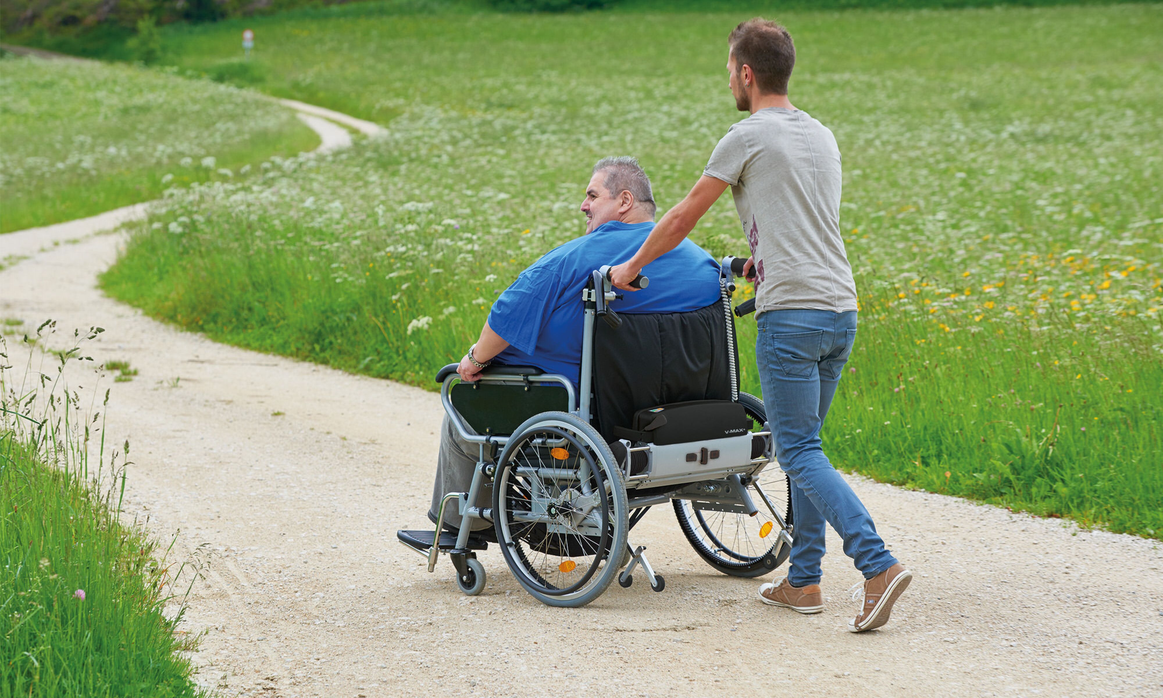 Das Bild zeigt einen sehr schlanken jungen Mann, der einen Mann mittleren Alters in einem sehr breiten Rollstuhl aufwandslos schiebt. Der Insasse nutzt aufgrund seines hohen Körpergewichtes einen Rollstuhl mit Zusatzantrieb V-MAX+, der speziell für adipöse Patienten entwickelt wurde. 