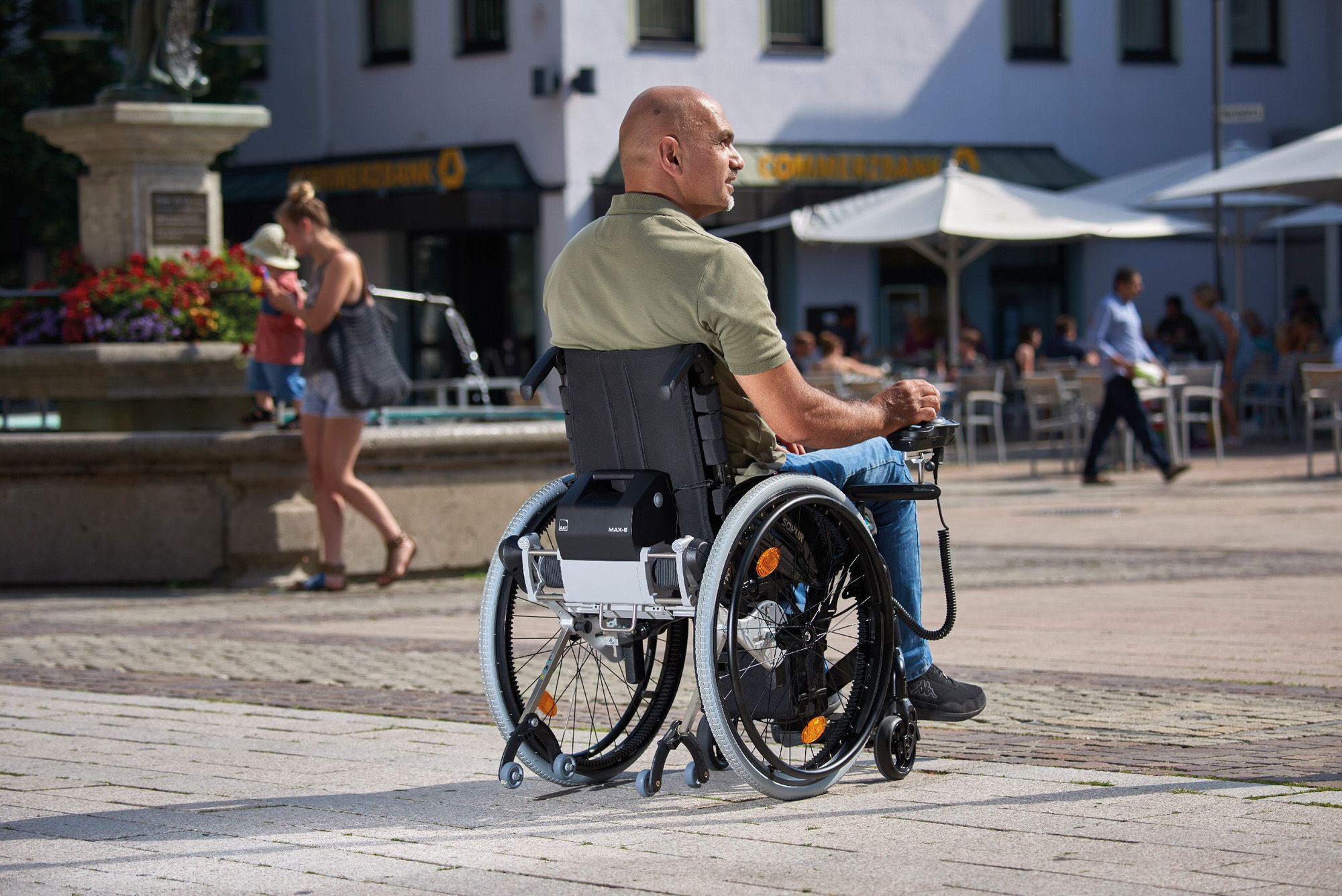 Das Bild zeigt einen Mann im Rollstuhl, der an einem sonnigen Tag einen Ausflug in die Innenstadt macht. Er nutzt einen Rollstuhl-Zusatzantrieb mit Joystick.