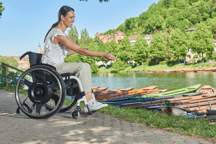 Auf dem Bild ist eine Frau mittleren Alters zu sehen, die in einem weißen Rollstuhl mit schwarzen Radnabenantrieb zur Restkraftverstärkung am Ufer eines Flusses sitzt und lächelnd Enten füttert. 