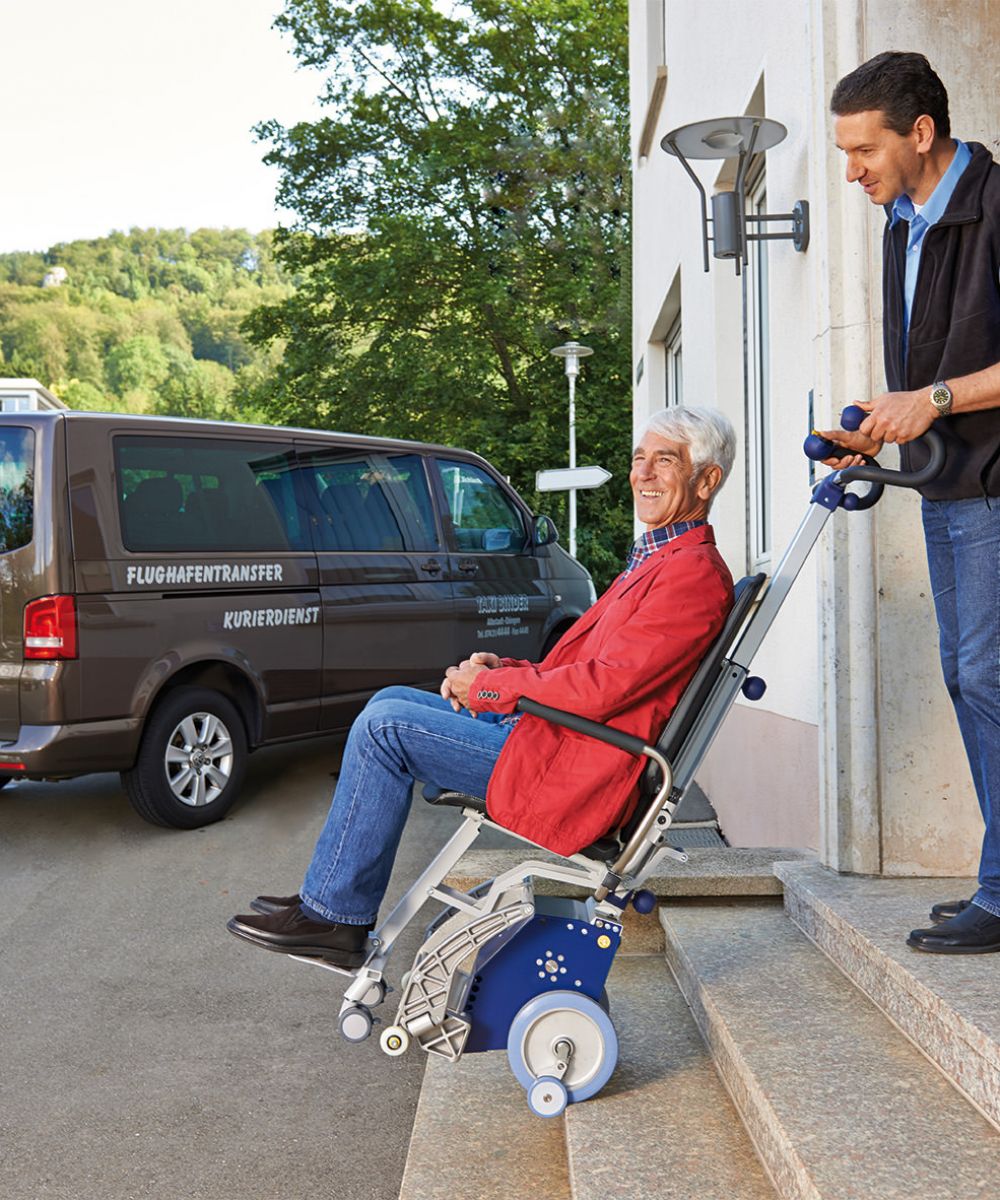 Das Bild zeigt einen Mann mittleren Alters, der einen älteren Mann, der in einem Treppensteiger mit integrierter Sitzeinheit sitzt, die Außentreppe eines Hauses hinunterbefördert. Im Hintergrund steht ein Fahrzeug für den Personentransport / Flughafentransport. 
