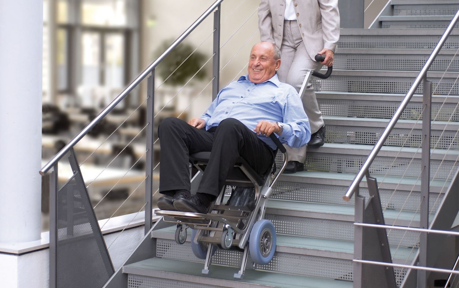 Das Bildzeigt einen Mann, der in seinem Rollstuhl sitzt und von einer Frau mit Hilfe eines angebauten Treppensteigers über eine Treppe transportiert wird. 