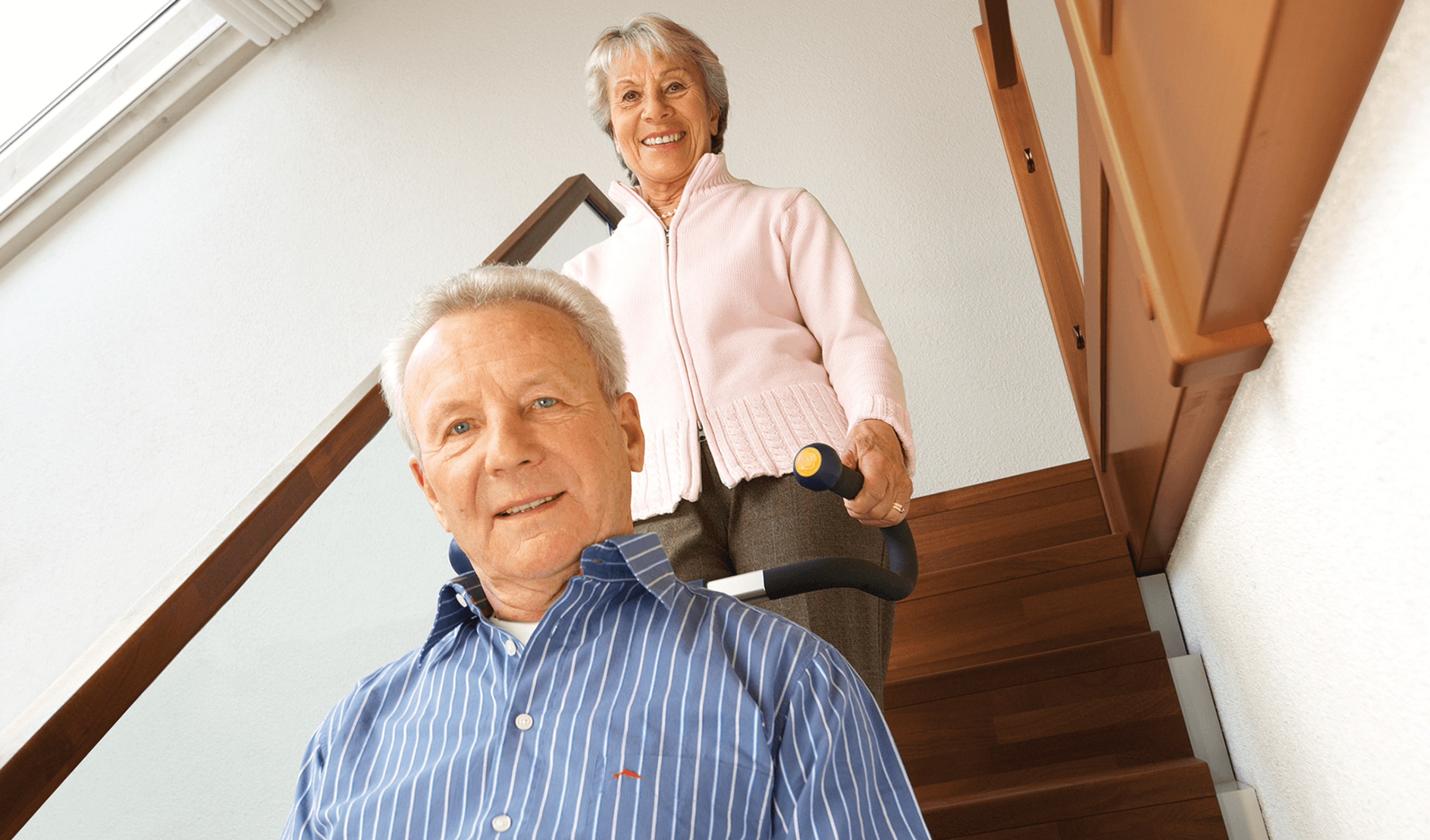 Das Bild zeigt einen Mann mittleren Alters, der von einer Frau in seinem Rollstuhl mithilfe eines Treppensteigers die Treppen hinaufbefördert wird. Beide lächeln in die Kamera.