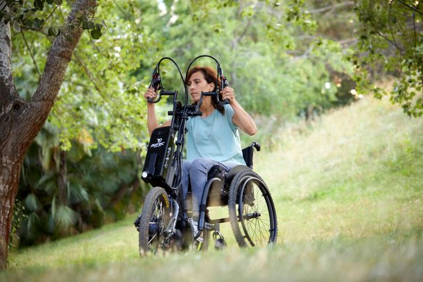 The picture shows a middle-aged woman who has attached a hybrid handbike to her wheelchair and is riding it through a forest. It can be seen that inclines and declines can also be mastered. The woman is operating the crank arms of the handbike with her hands. 