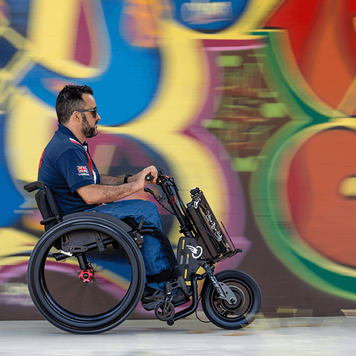 Das Bild zeigt einen jüngeren Mann mit Sonnenbrille, der mit einem Zuggerät MINI 2 von BATEC, welches an seinem schwarz roten Rollstuhl angebaut ist, schnell vor einer bunten Graffitiwand vorbei fährt.  