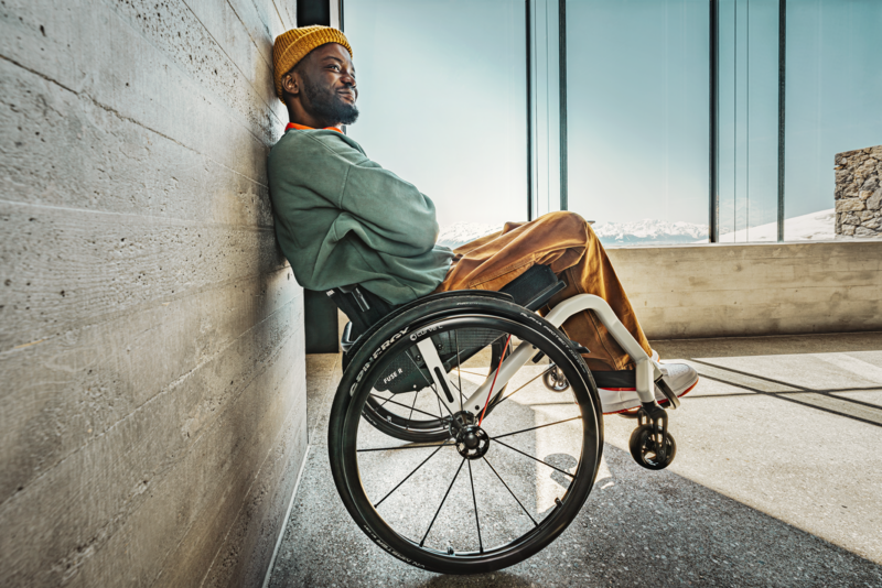 The picture shows a young man in a FUSE-R active wheelchair leaning back against a wall. A white mountain backdrop can be seen in the background. 