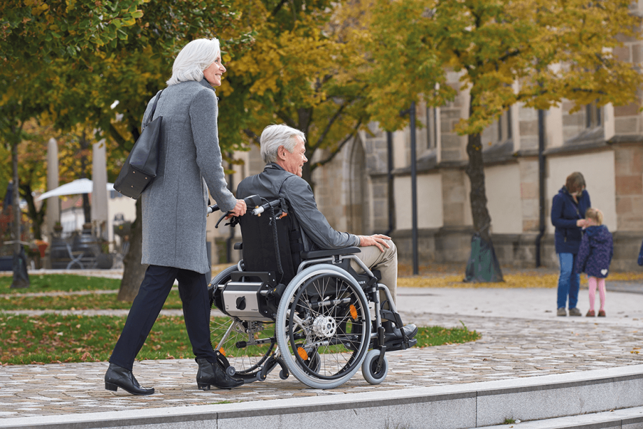 Das Bild zeigt eine ältere Frau, die einen älteren Mann im Rollstuhl über einen gepflasterten Gehweg schiebt. Beide lächeln und passieren eine grüne Wiese mit einer Kirche im Hintergrund. Am Rollstuhl befindet sich die Schiebe- und Bremshilfe V-MAX2, welche die Frau beim Schieben und Abbremsen des Rollstuhls unterstützt.