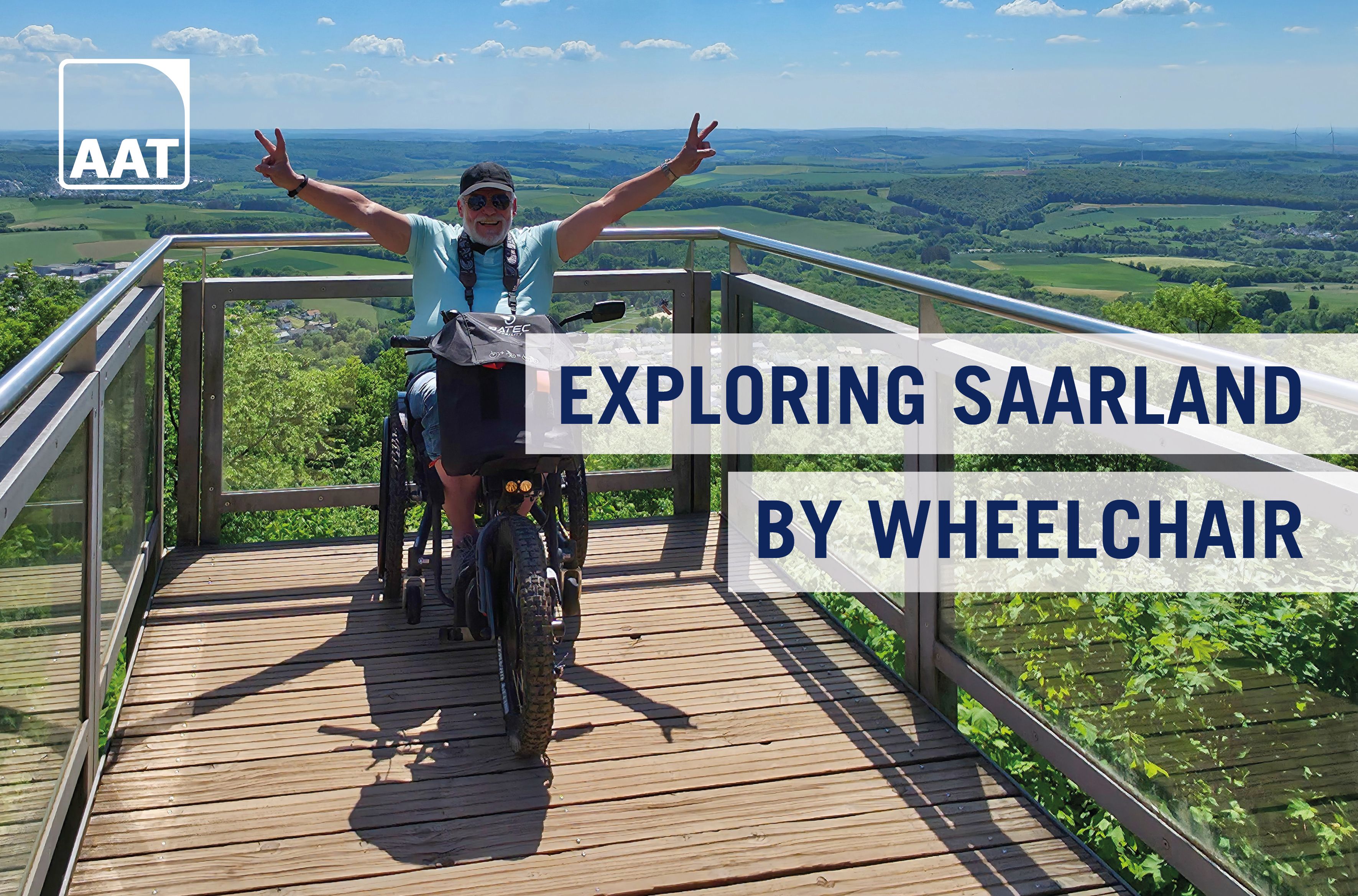The picture shows Hans-Jürgen Rohe standing on a viewing platform above the Saarland with his SCRAMBLER 2 wheelchair and wheelchair traction device from BATEC and AAT. He stretches his hands in the air and shows two peace signs with his fingers.