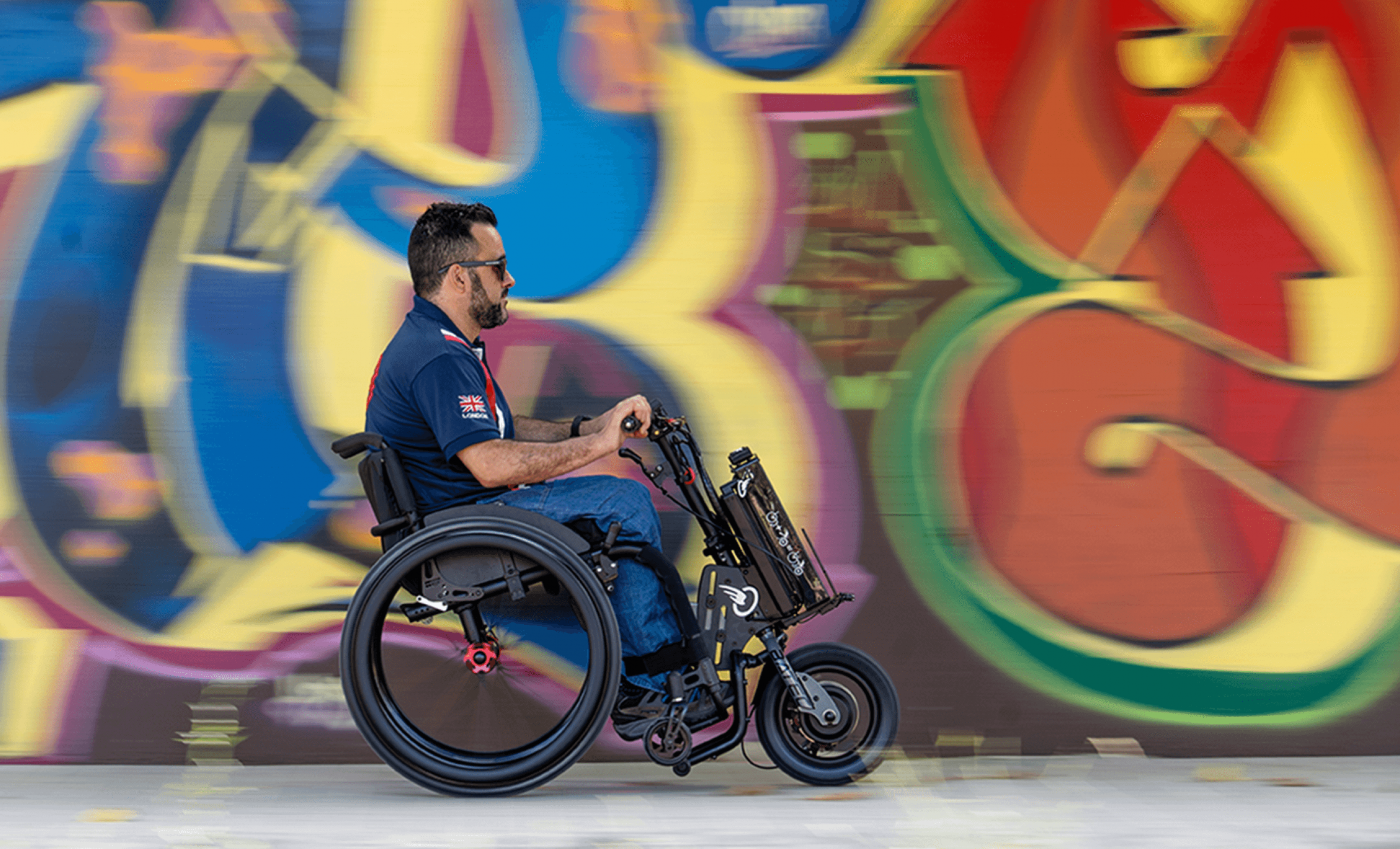 Das Bild zeigt einen jüngeren Mann mit Sonnenbrille, der mit einem Zuggerät MINI 2 von BATEC, welches an seinem schwarz roten Rollstuhl angebaut ist, schnell vor einer bunten Graffitiwand vorbei fährt.  