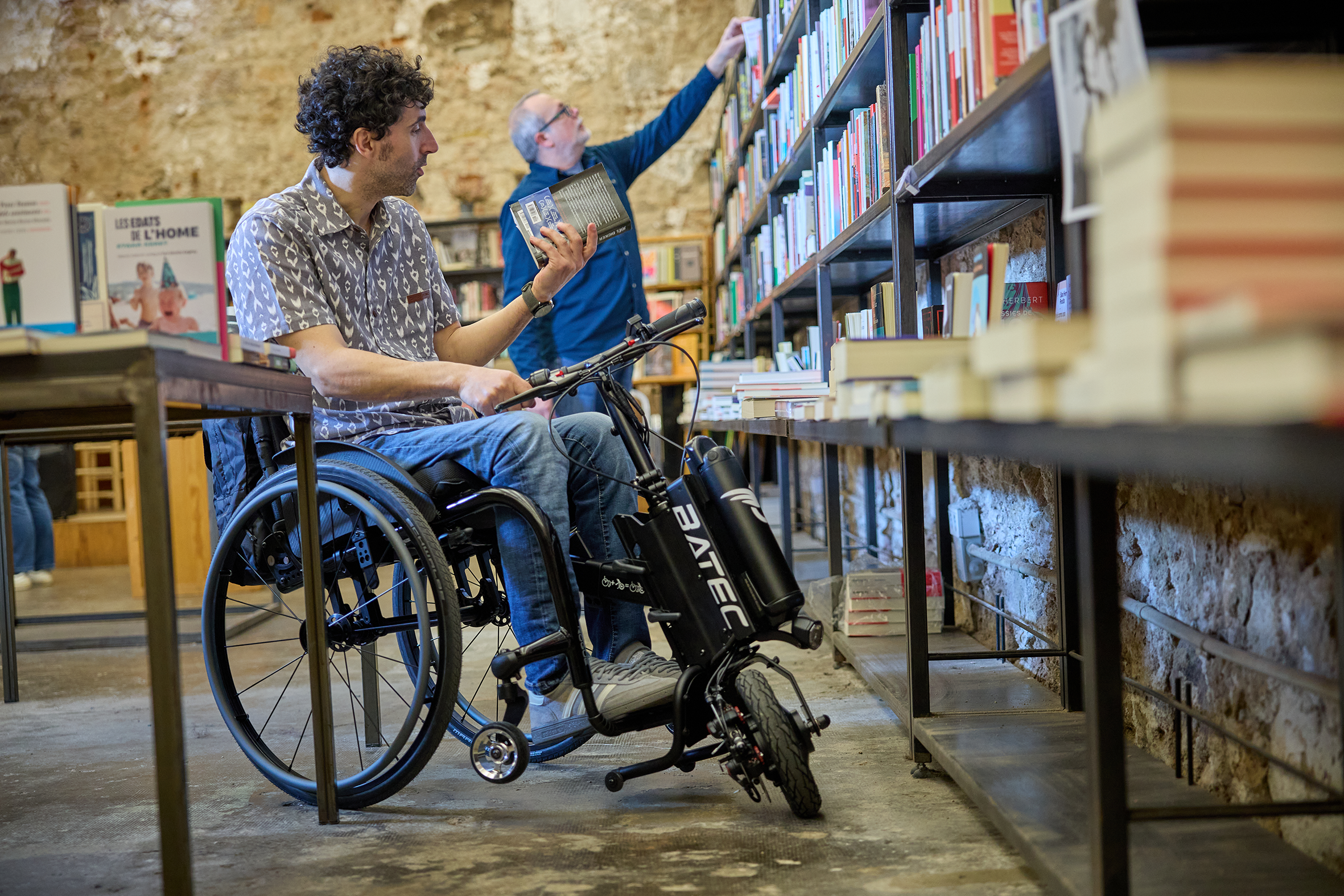 The picture shows a young man standing in a cramped space in a ventriloquist's shop with his manual wheelchair and BATEC Air traction device.