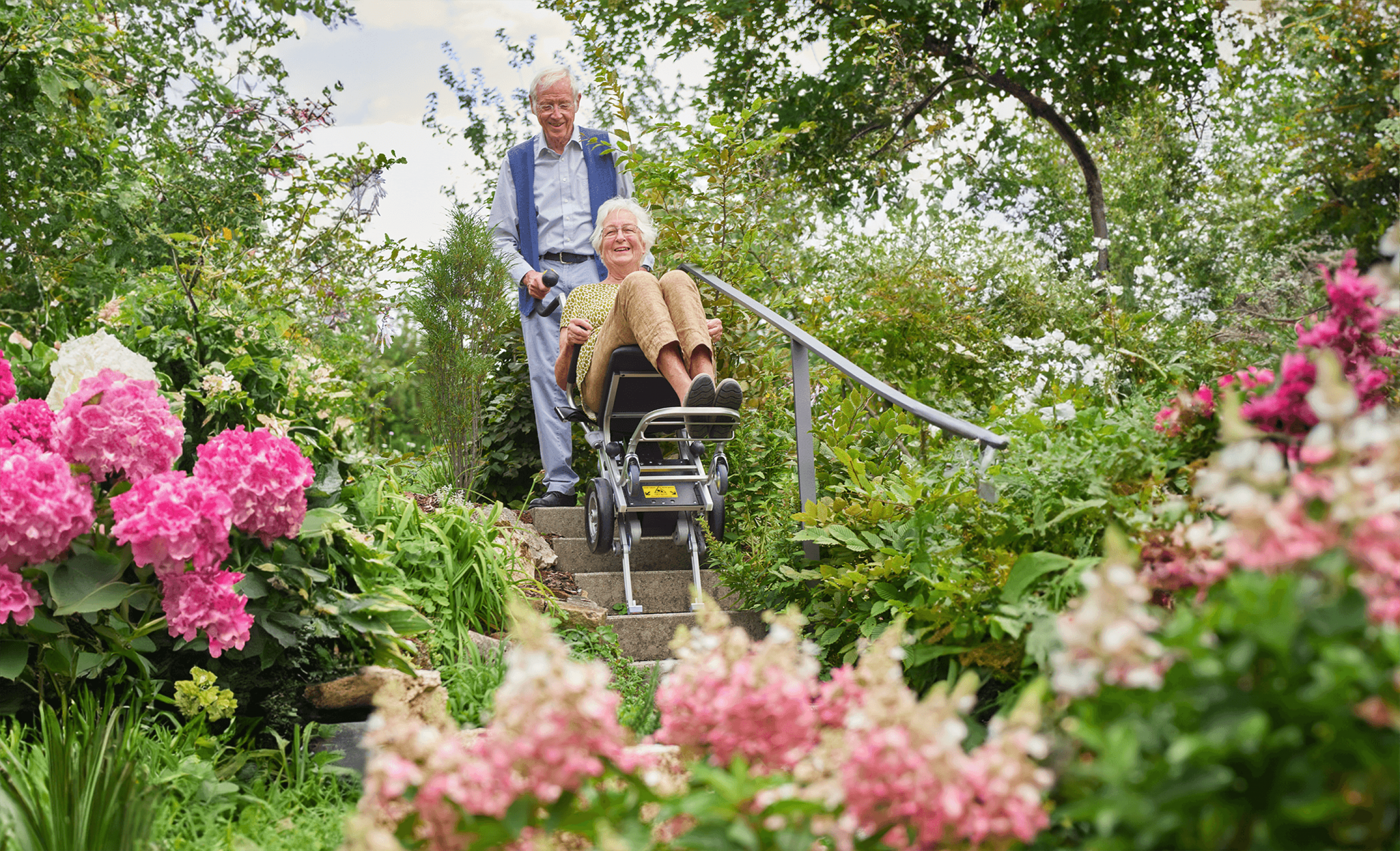 Das Bild zeigt einen älteren Mann, der seine Frau zu Hause im Garten die Treppen hinaufbefördert. 