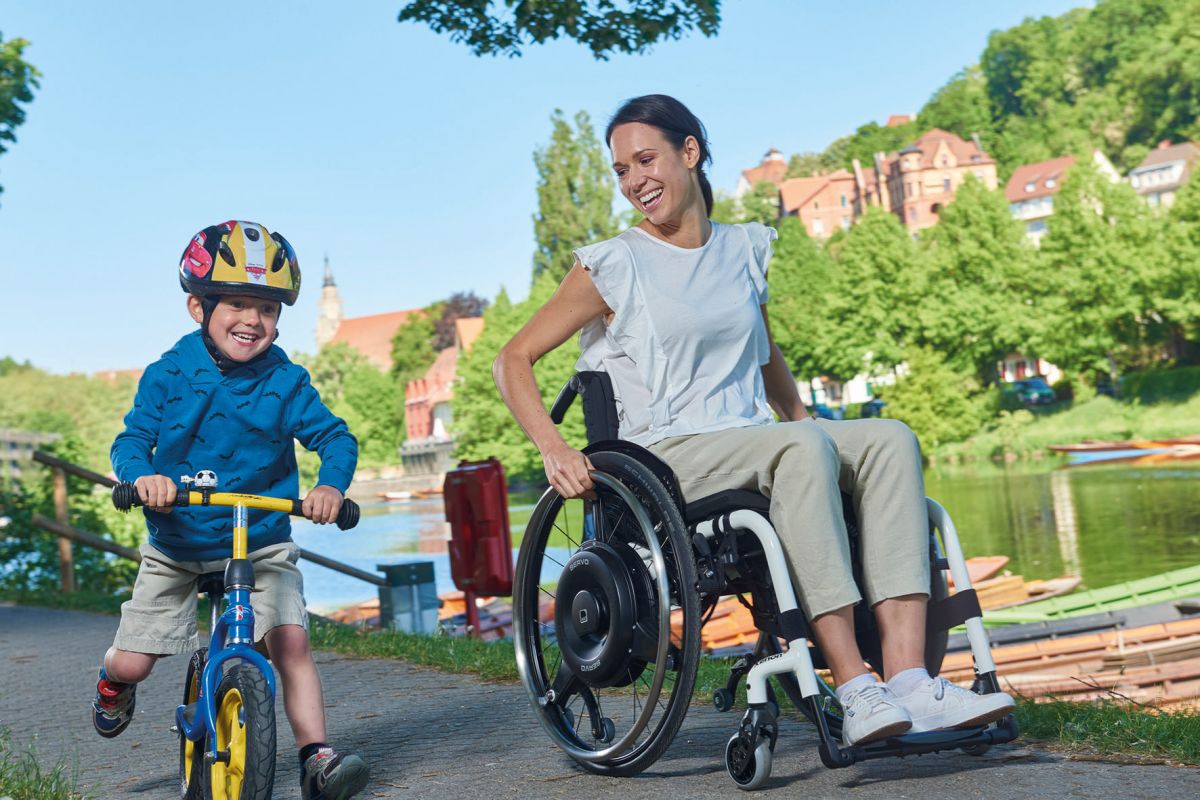 The picture shows an elderly man in a wheelchair being pushed by a young woman. There is a V-MAX mini push aid for wheelchairs on the wheelchair, which is helping the woman to push.
