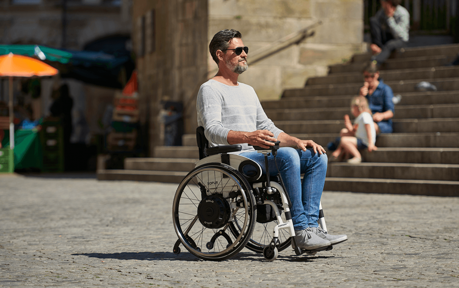 The picture shows a middle-aged man driving his wheelchair through a sunny city center. He is driving over cobblestones and steering his wheelchair himself using a joystick. The wheelchair wheels are fitted with a SOLO wheel hub drive that powers the wheels. 